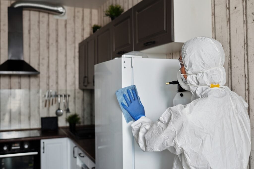 A person cleaning a kitchen in a protective suit, wearing a dust mask and eye protection