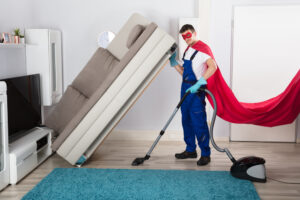 man in cape deep-cleaning lifting couch with vacuum-cleaner in living room