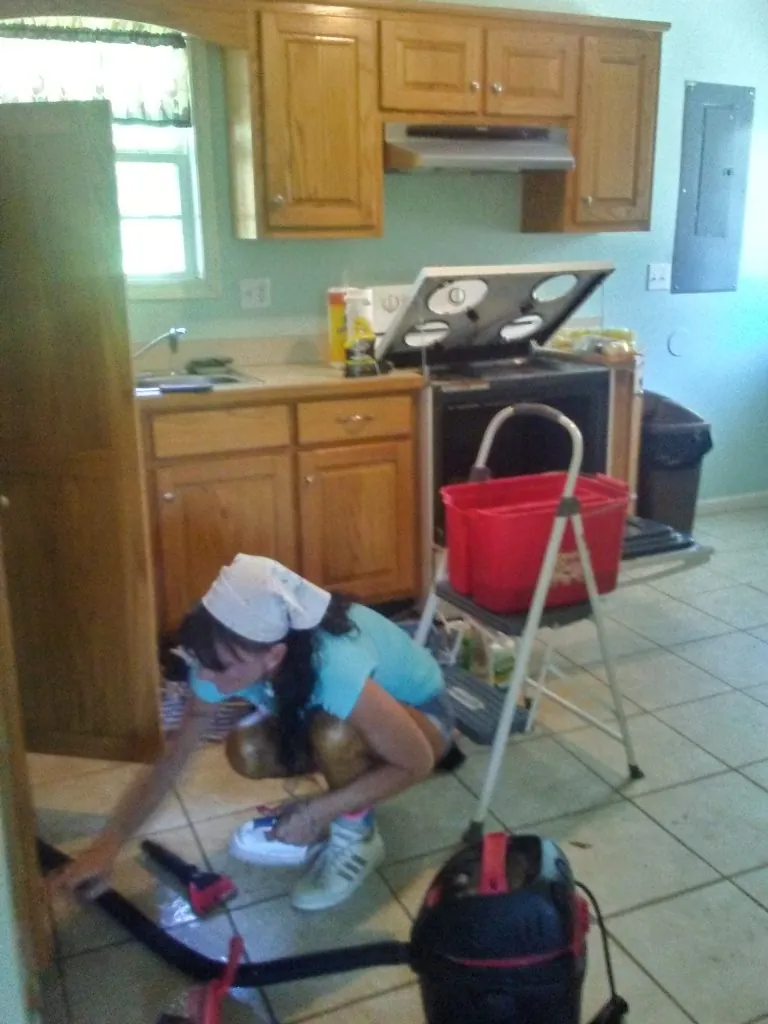 Tabby cleaning a kitchen floor beside an open lower cabinet.